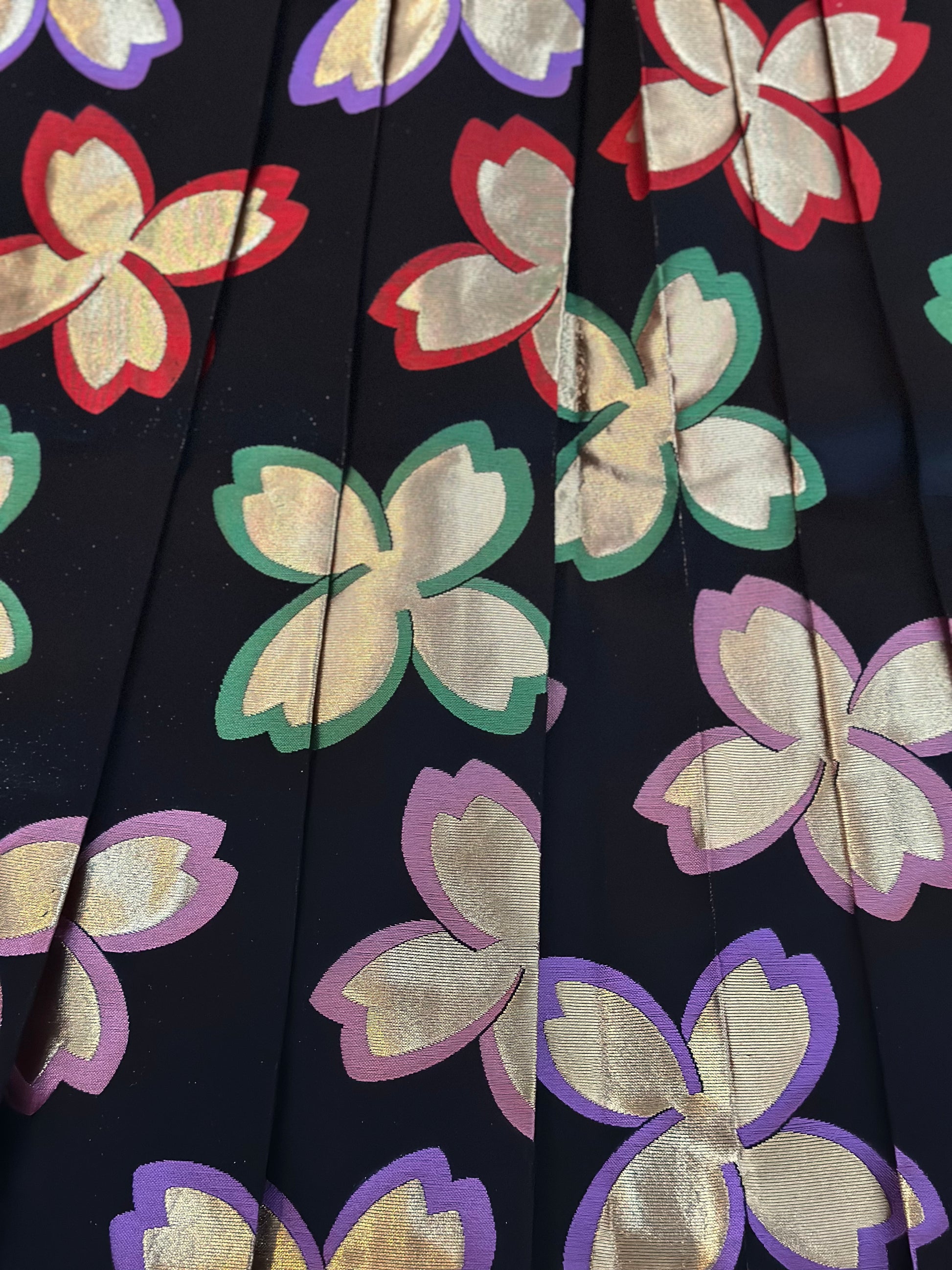 Close-up of a pleated fabric surface with a repeating floral pattern in metallic gold, red, green, and purple on a very dark  black ground.