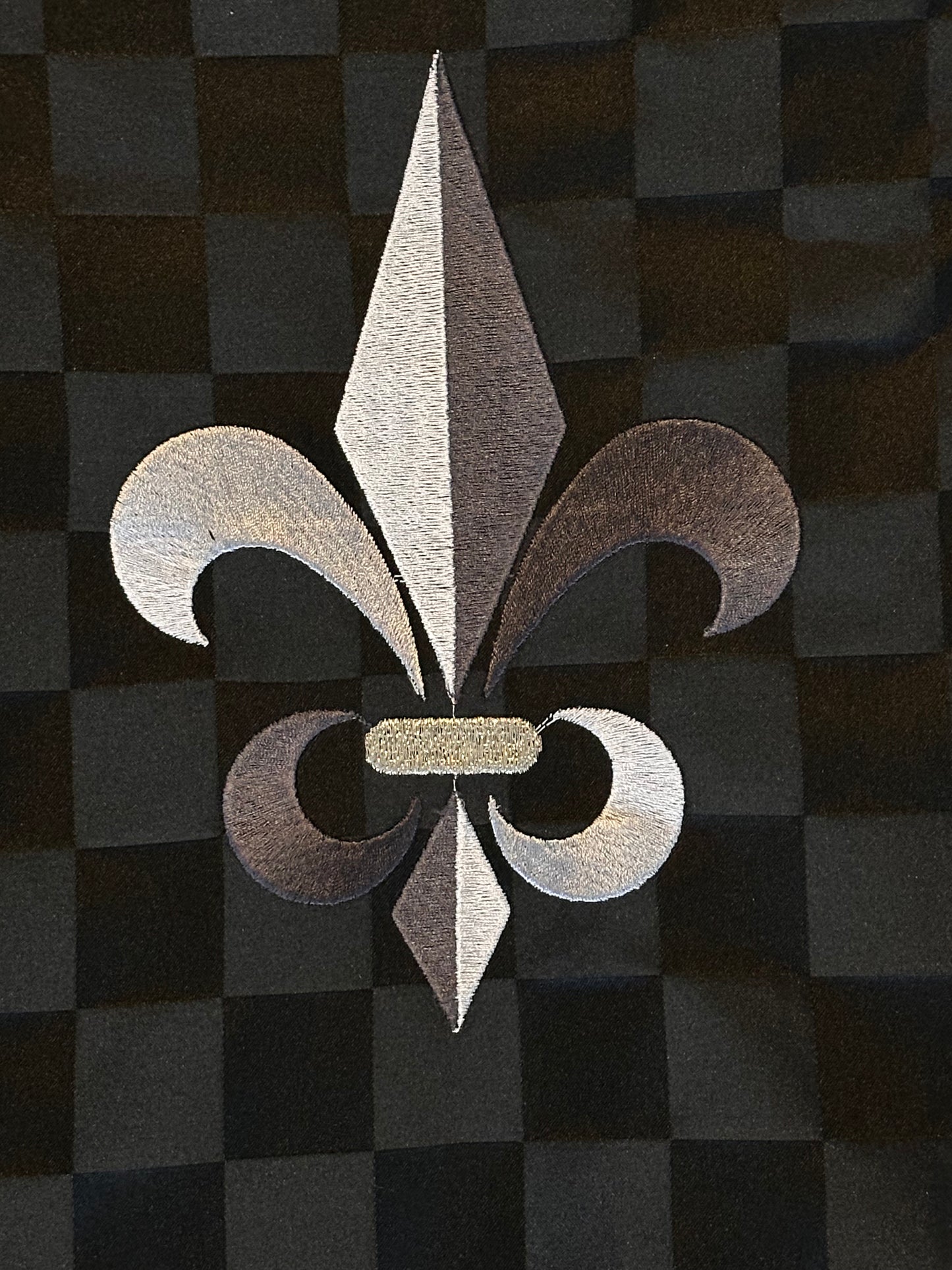 A close up of a metallic silver fleur-de-lis emblem centered on a black and dark gray checkered background.