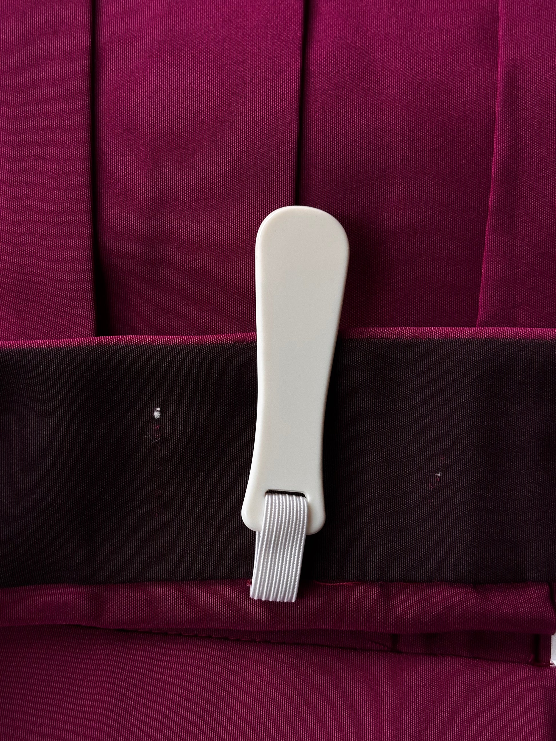 A close-up of a white plastic garment clip holding a dark charcoal fabric band over deep magenta-purple pleated fabric.