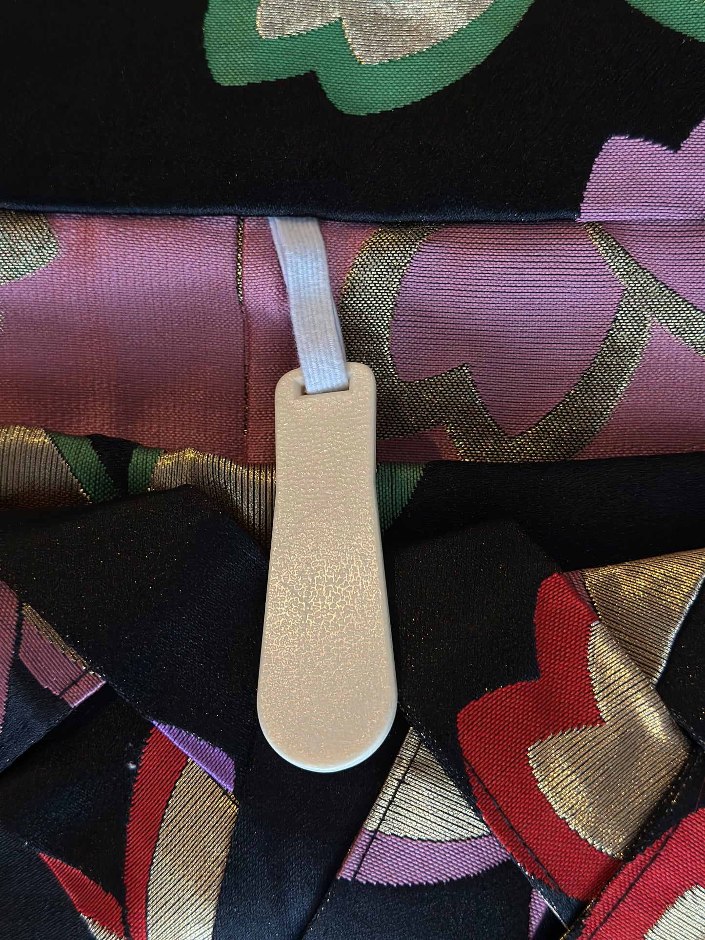 A close-up of a folded garment featuring a black base with a vibrant, multi-colored pattern of pink, red, green, and gold. A light beige plastic strap is attached with a white elastic band.