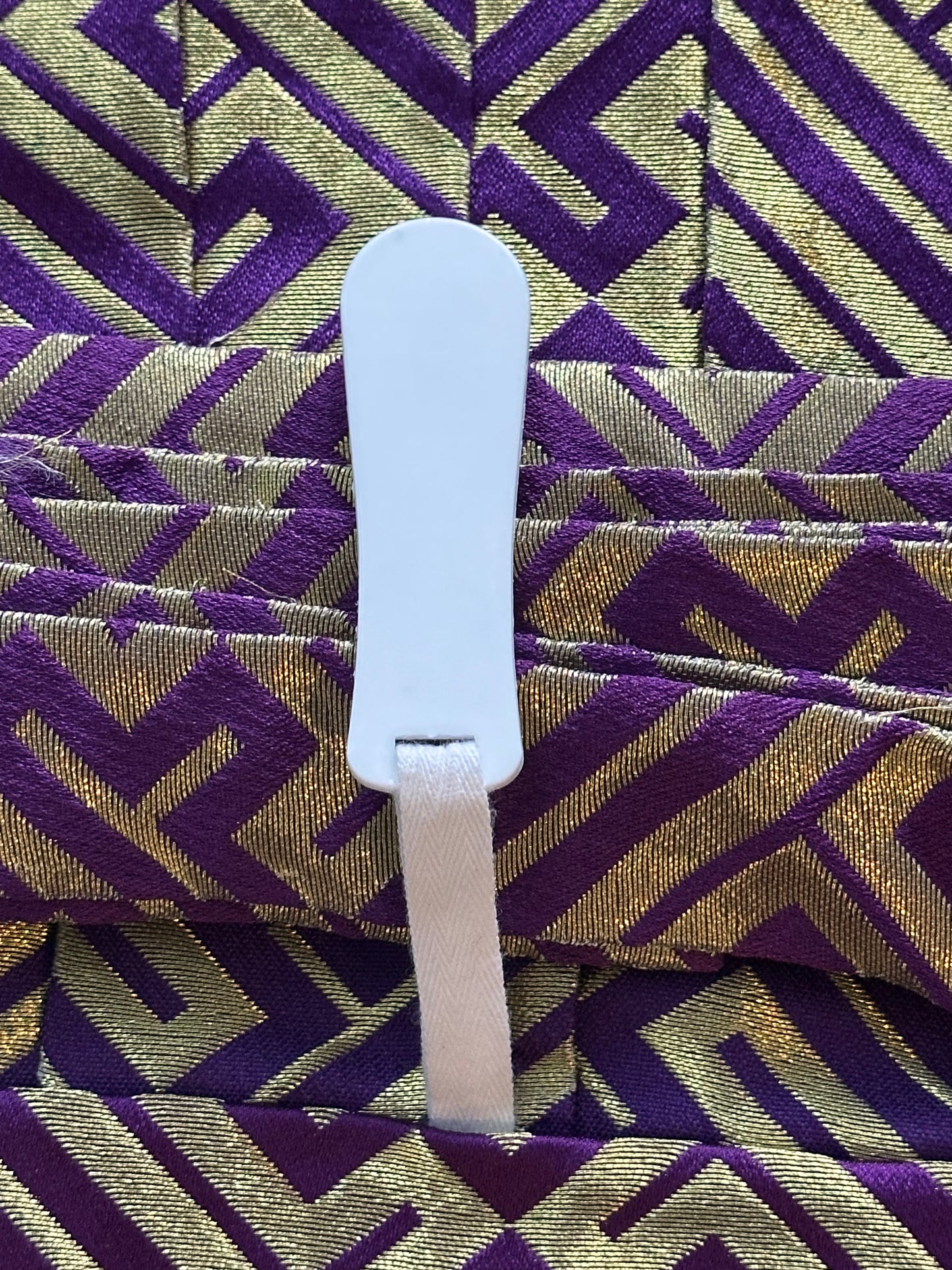 A close-up of a white plastic and fabric strap component attached to a purple and gold patterned textile background.