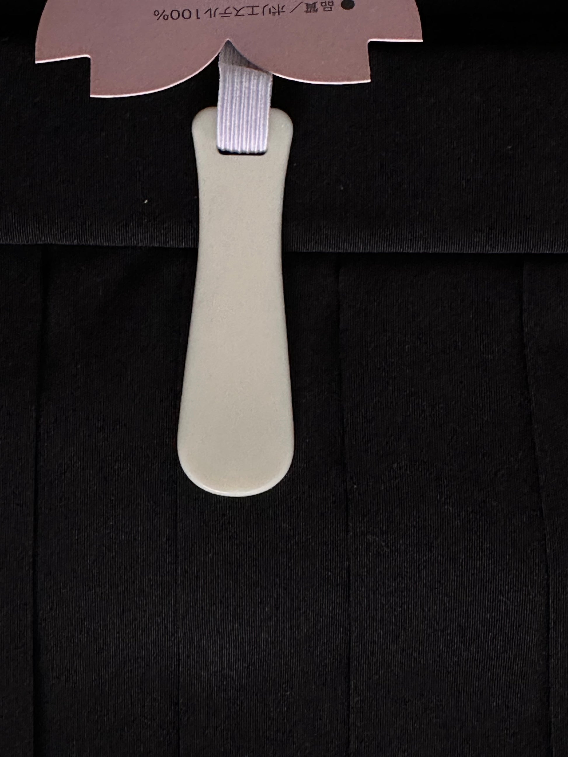 A close-up of a black hakama fabric, featuring a light beige plastic hanger insert with a lavender elastic strap and a pink paper tag at the top.