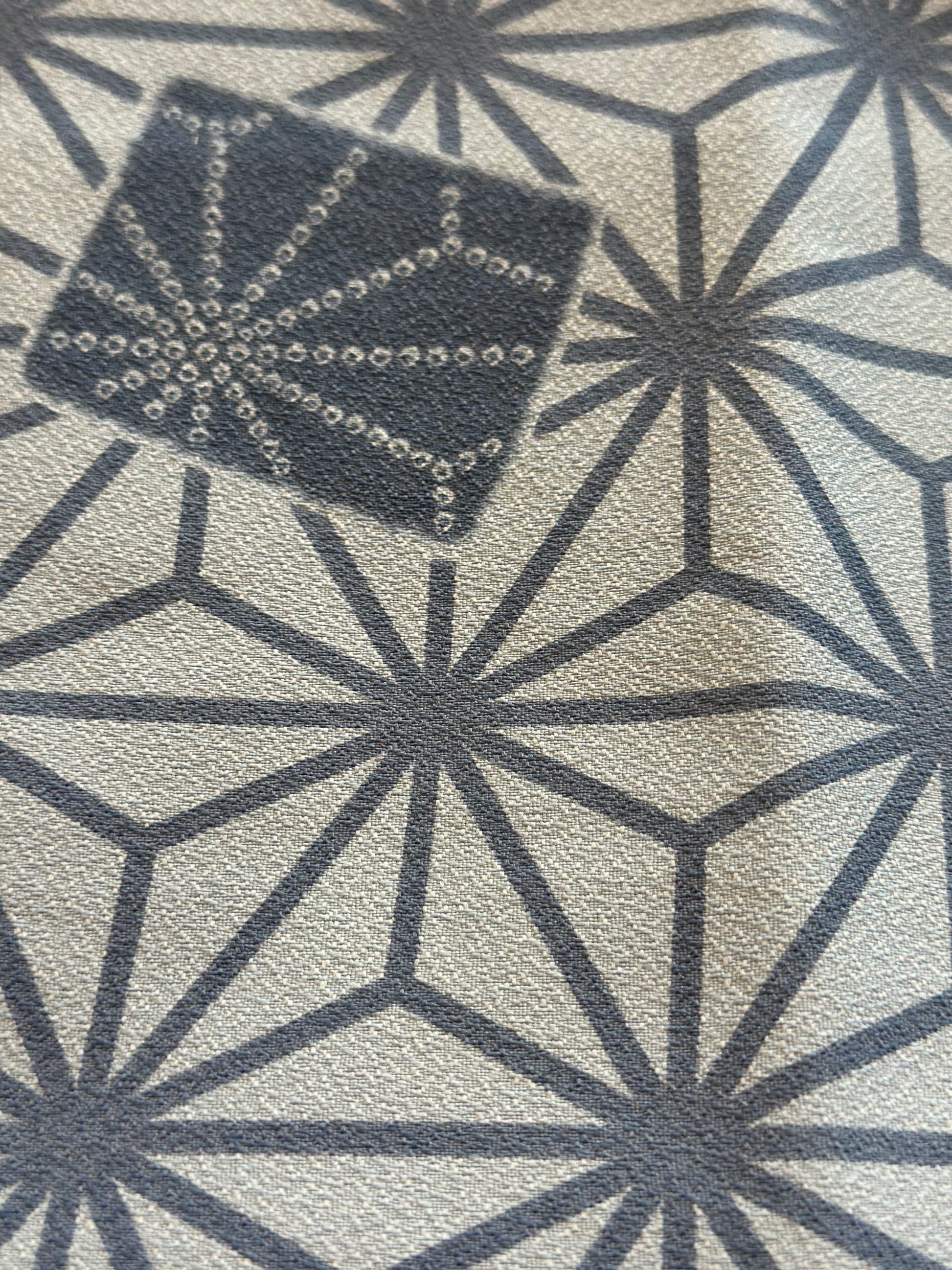 A close-up of a textured rug featuring a dark gray geometric starburst pattern on a light beige background, with a small inset showing a matching dark gray textured sample piece.
