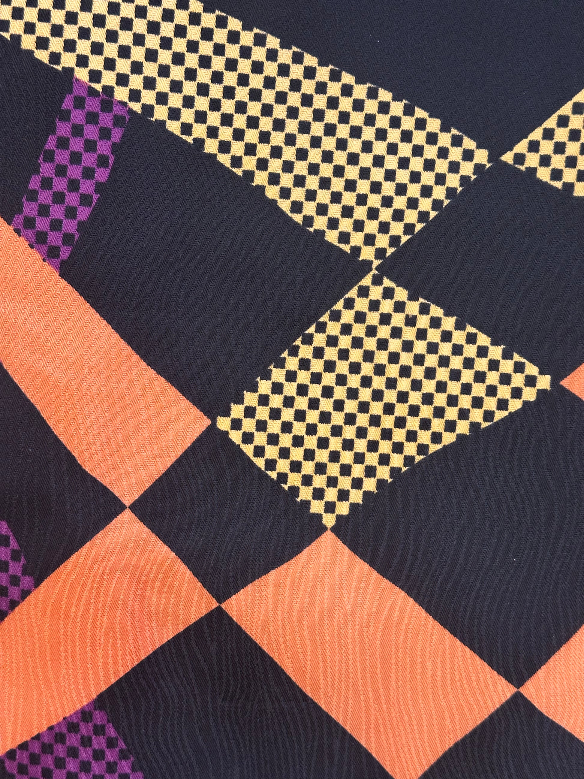 A close-up of a geometric patterned fabric featuring diagonal stripes and diamond blocks in orange, yellow-and-black checkered pattern, and purple-and-black checkered pattern on a dark navy background.
