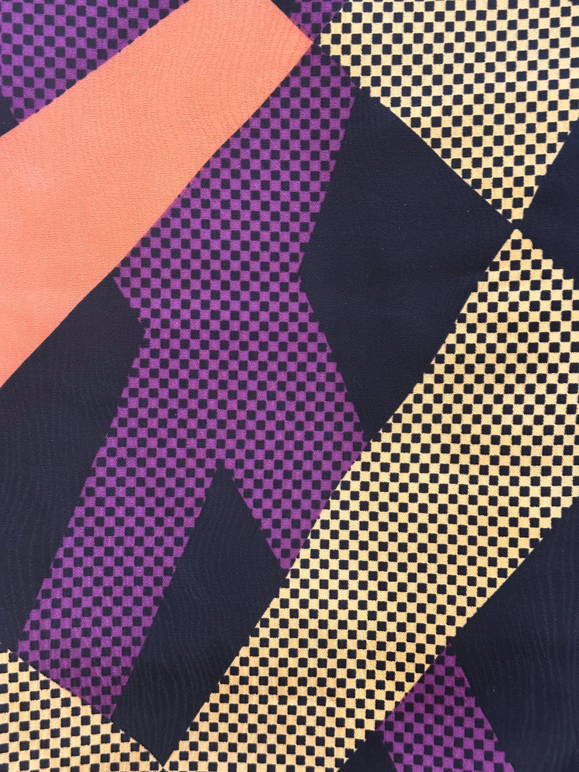A close-up of a geometric patterned textile featuring diagonal bands of purple, peach, navy, and beige with black checkered and dotted textures.