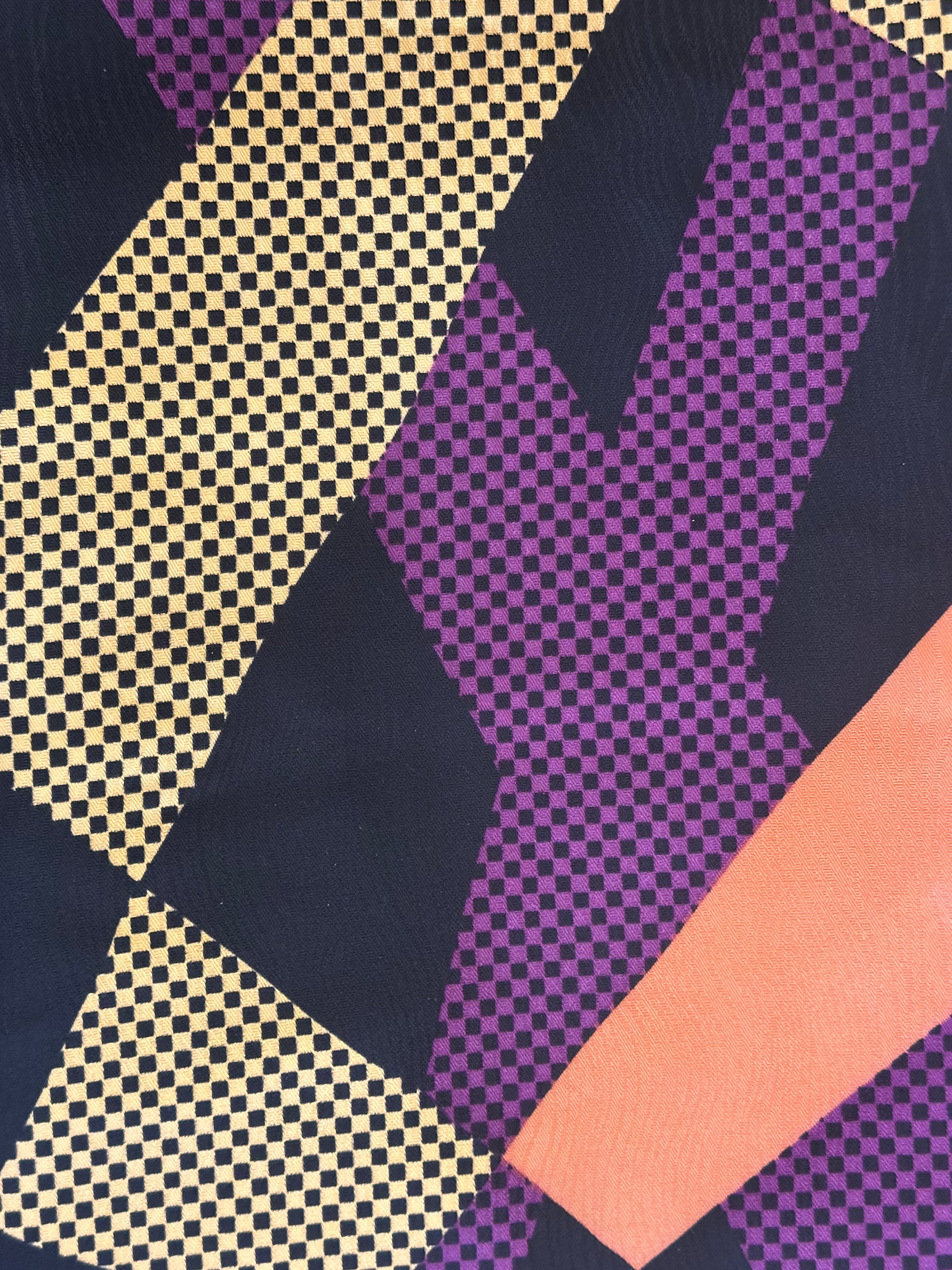 A close-up of a patterned textile with large geometric blocks in purple, navy, peach, and beige, including checkerboard and dotted textures.