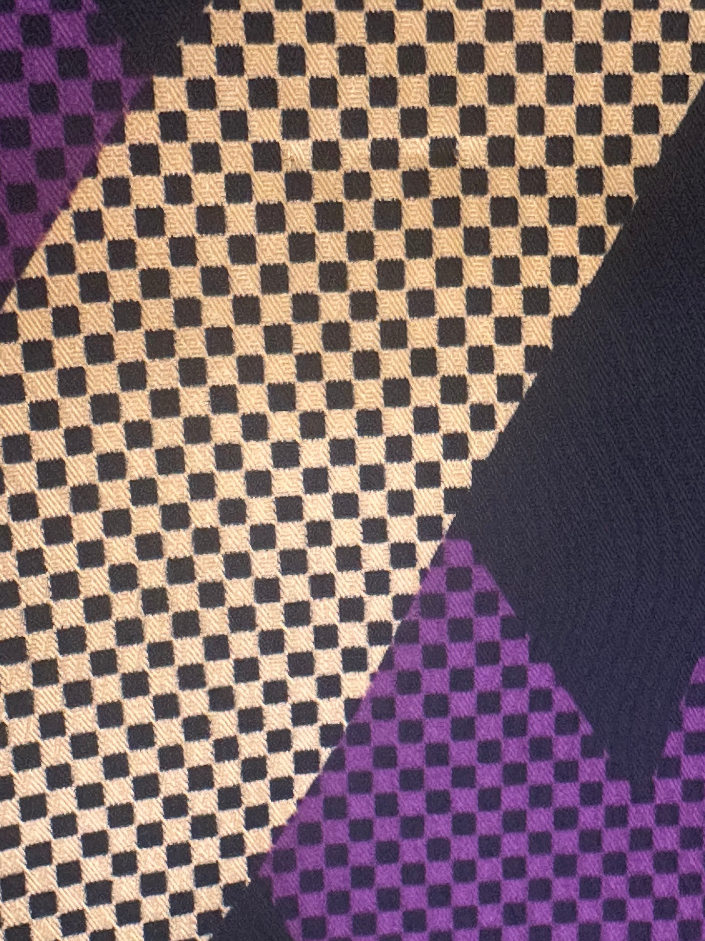 A close-up of a patterned textile featuring a beige and black checkered design with purple geometric sections and a dark navy diagonal band.