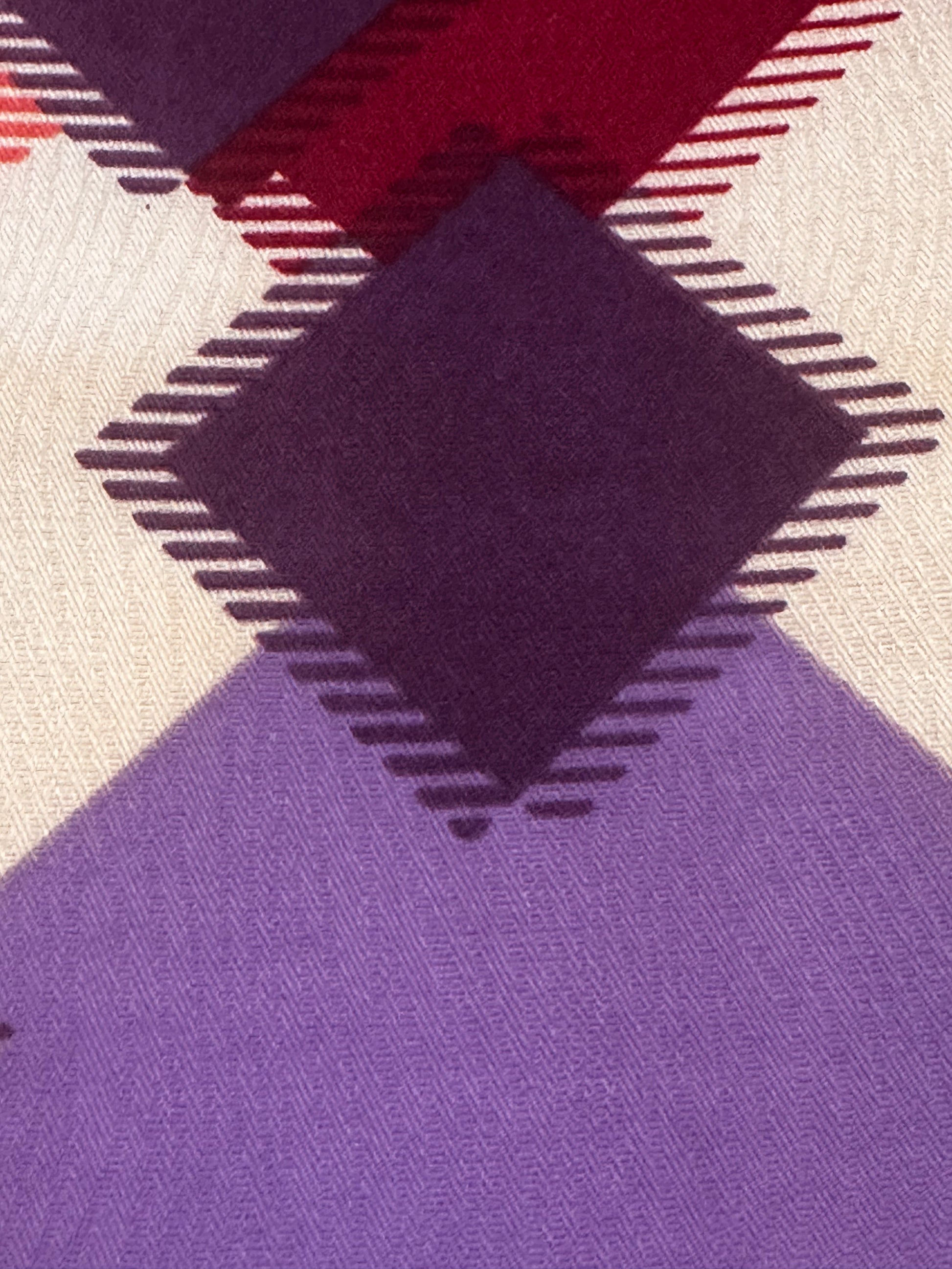 A close-up of a woven textile pattern with large diamond and triangular sections in purple, cream, burgundy red, and dark plum, outlined with dashed stitching.